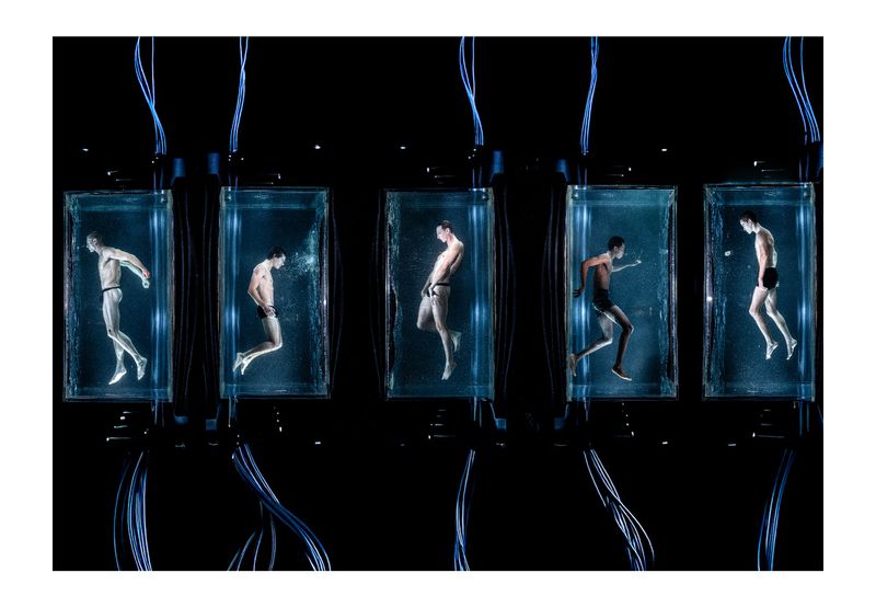 Five adult males in swim briefs are submerged in glass tanks with water, cables, and blue lighting against a dark background.