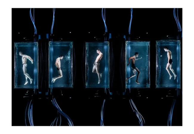 Five adult males in swim briefs are submerged in glass tanks with water, cables, and blue lighting against a dark background.