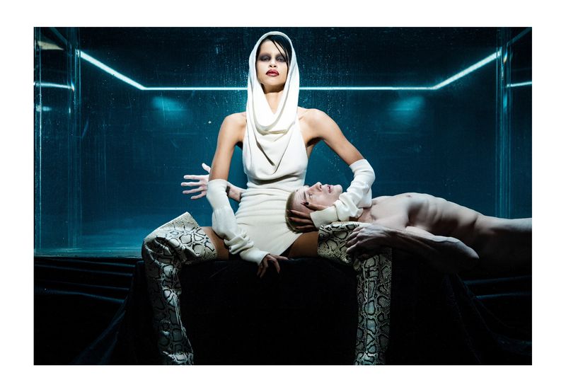 A model in a white dress and head covering sits astride a shirtless male model lying on a black surface in a studio with blue lighting.