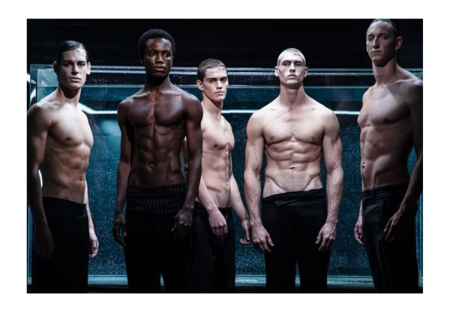 Group of five shirtless, fit men in black pants strike poses against dark backdrop and lit glass wall for a fashion or fitness editorial.