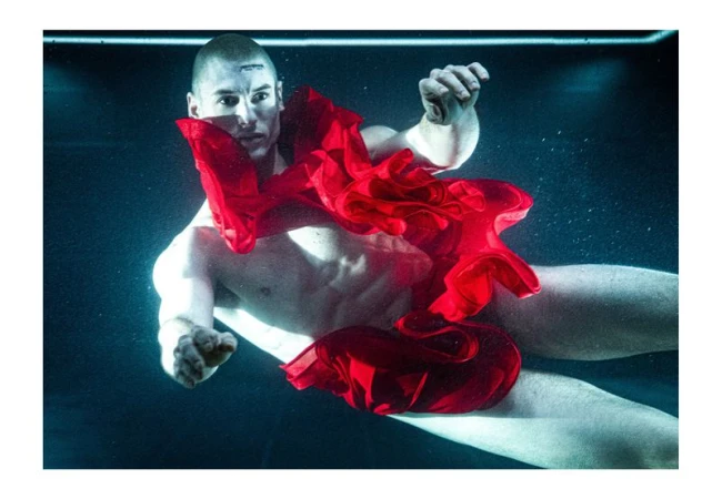 Underwater portrait of a muscular adult male with a shaved head, partially nude, adorned with red fabric in an ethereal, aquatic setting.
