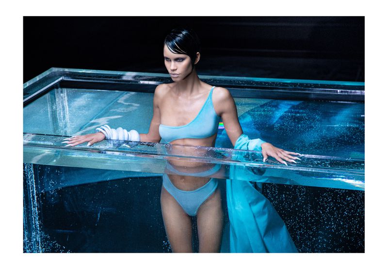 Brunette woman in a light-blue bikini and matching robe stands in a water-filled glass box in an indoor environment with dark background.