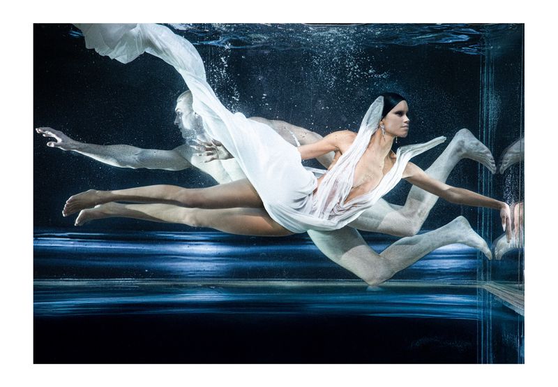 Underwater fashion photography with a woman in a flowing white dress and a man swimming in the background with blue lighting.