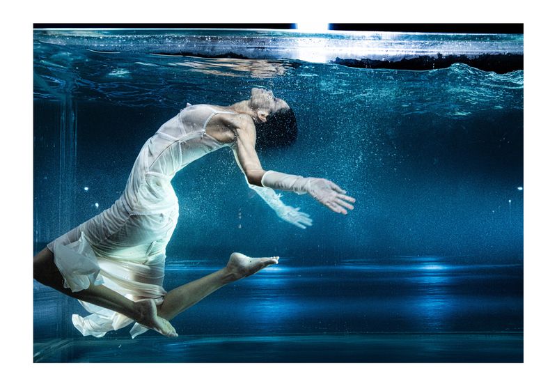 Woman in a white dress underwater in a pool, gracefully suspended with her arms outstretched, creating a serene, ethereal scene.