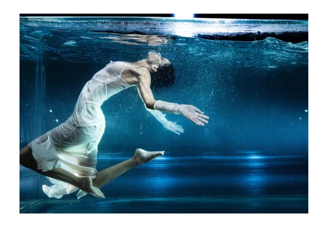 Woman in a white dress underwater in a pool, gracefully suspended with her arms outstretched, creating a serene, ethereal scene.