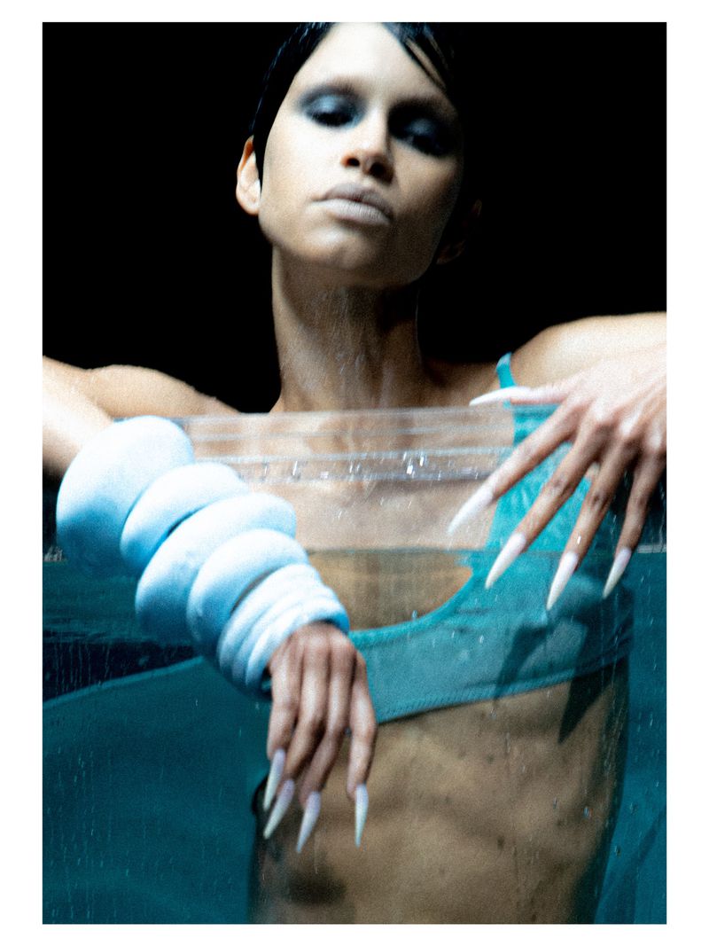 Dramatic studio portrait of a woman partially submerged in water, wearing avant-garde aqua fashion and long stiletto nails against a black backdrop.