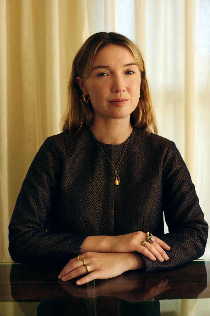 Portrait of a woman with fair skin and light hair, wearing a dark blazer and jewelry, sitting at a table indoors with neutral curtains.