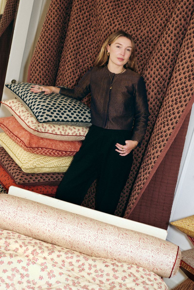 A woman poses indoors amid stacked patterned pillows and fabric rolls, showcasing textile designs in a warm, inviting setting.