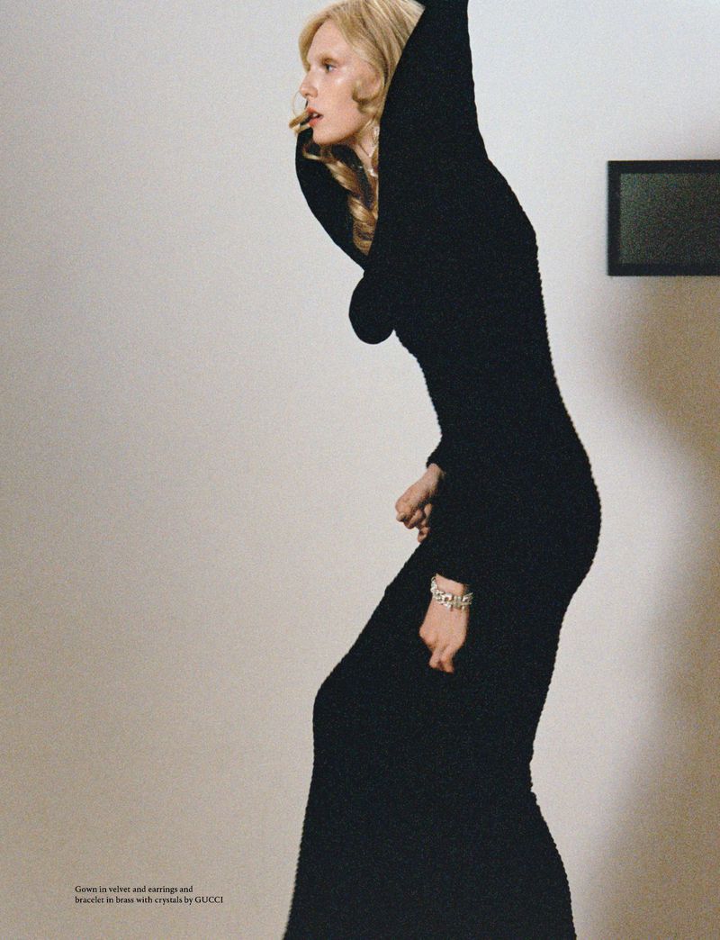 Blonde woman in a black hooded dress with a cigarette in her mouth poses in front of a white wall with a black-framed painting.