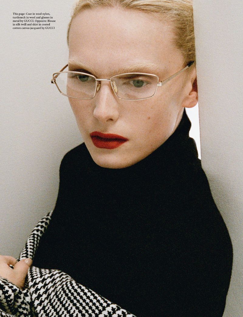 Close-up of a woman with blonde hair and red lipstick wearing glasses and a black turtleneck with a houndstooth jacket over her shoulder.