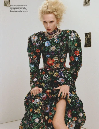 A blonde model wearing a black gown with colorful sequined floral embroidery and a statement necklace against a minimalist white wall.