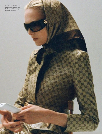 A fashionable woman wearing sunglasses and a headscarf with a patterned jacket, looking at a smartphone in an indoor setting.