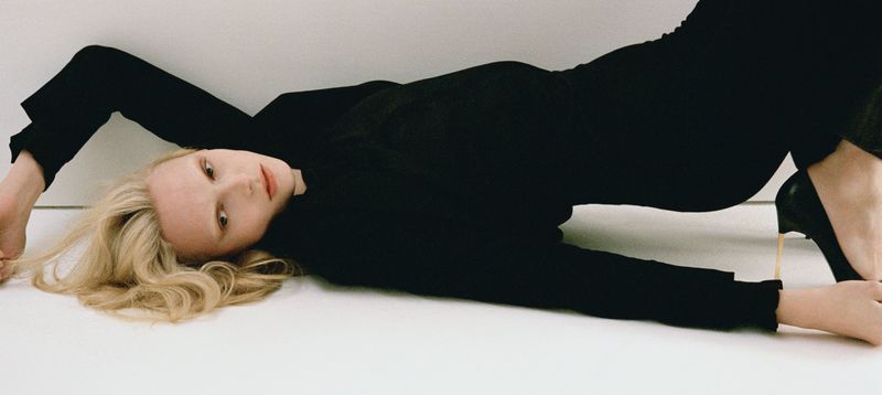 Blonde woman in black clothing poses on a white surface, wearing high heels. Fashion photography, eye-level angle, studio lighting.