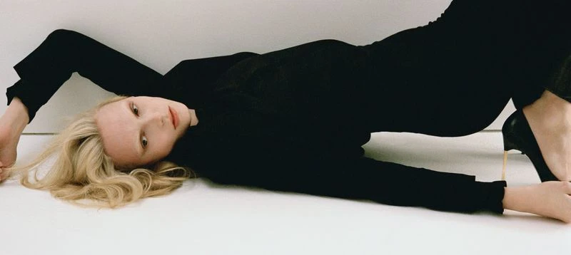 Blonde woman in black clothing poses on a white surface, wearing high heels. Fashion photography, eye-level angle, studio lighting.