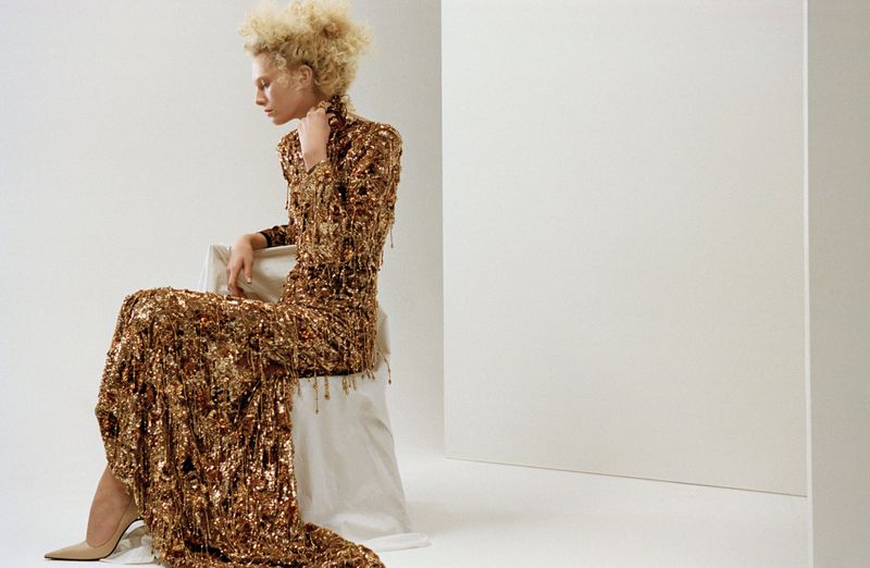 Fashion shot of a woman with blonde afro hair sitting in a gold sequin dress and heels in a white studio. Neutral, elegant fashion.