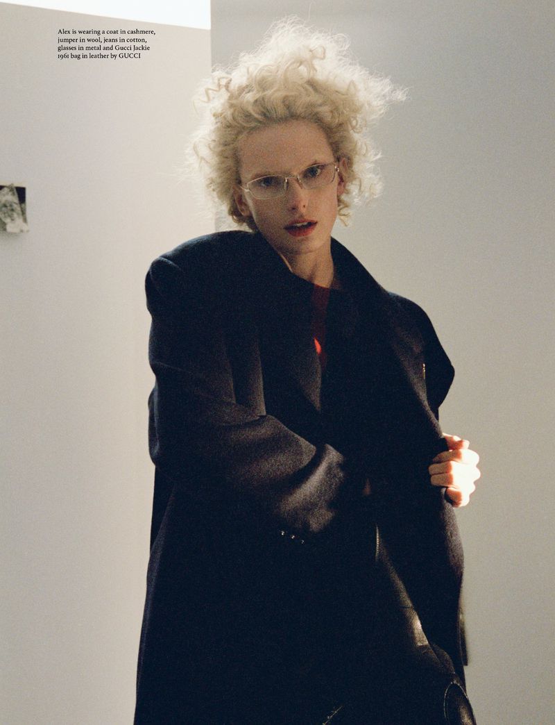 Fashion shot of a blonde adult posing in a coat and glasses indoors against a white wall with geometric artwork in the background.