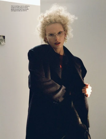 Fashion shot of a blonde adult posing in a coat and glasses indoors against a white wall with geometric artwork in the background.