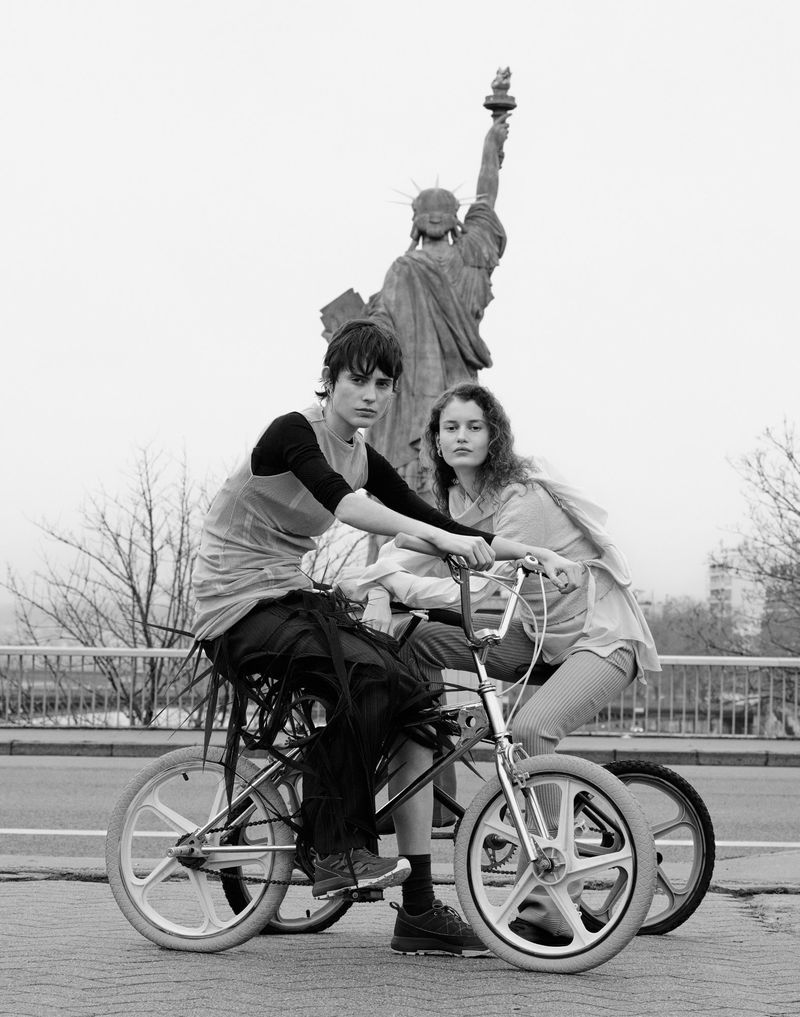 Two young adults posing with BMX bicycles on a city street, Statue of Liberty replica behind, monochrome, overcast sky.