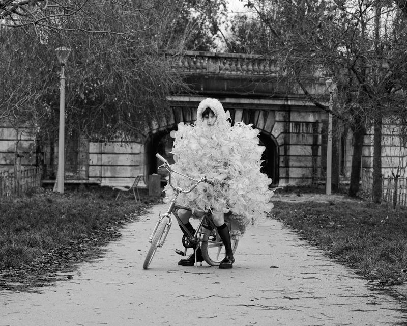 Black and white portrait of a person in an elaborate tulle costume riding a bicycle on a pathway with an arched bridge in background.