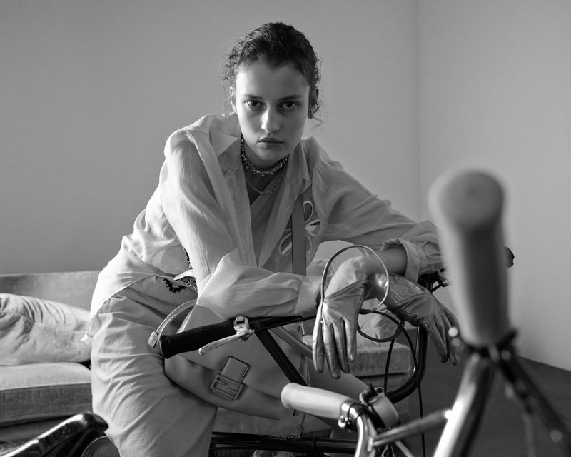 Black and white portrait of a young woman with a serious expression sitting on a bicycle in an indoor setting with soft lighting.