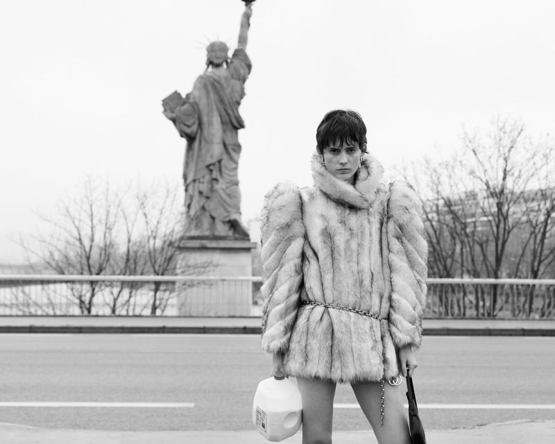 A person in a fur coat stands in front of the Statue of Liberty, holding a milk carton and a bag in a black and white fashion shot.
