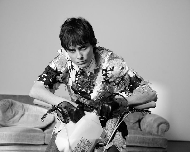 Monochrome portrait of a person with short hair leaning on a white motor scooter, wearing a floral top, with a grey couch in background.
