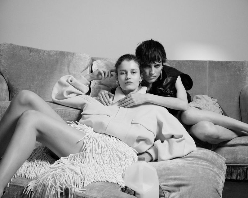 Two women pose on a gray couch in a black-and-white portrait. One wears a jacket with fringe. The other wears a black top.