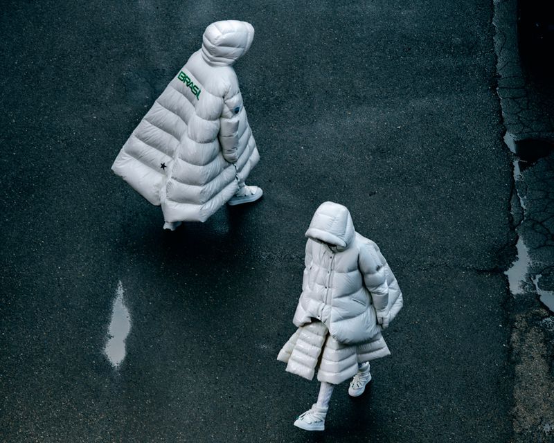 Overhead shot of two people wearing puffy white coats and sneakers walking on a textured grey surface with the word BRASIL on a coat.