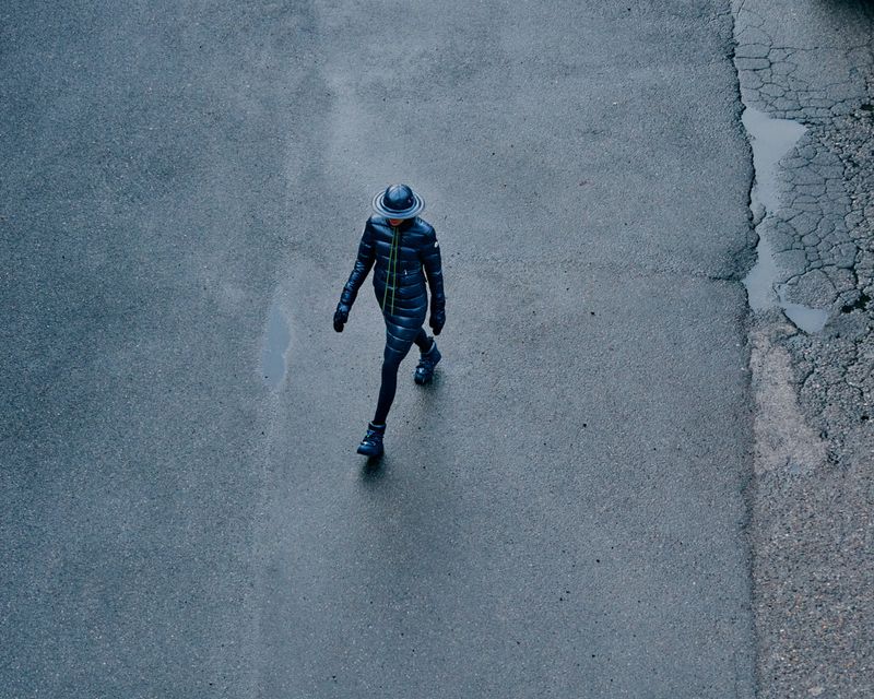 High-angle shot of a person walking on gray asphalt, wearing a dark puffer coat, matching pants, gloves, and a distinctive hat.