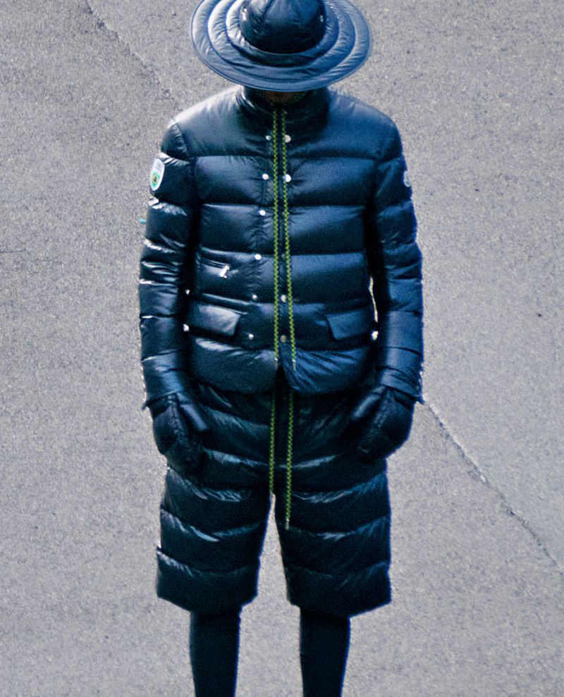 Model wearing dark quilted puffer jacket, matching shorts, gloves, and wide-brimmed hat standing on concrete.