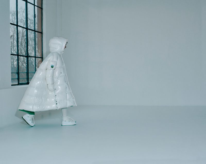 Person in a white puffer coat with hood walking in an empty white room with a window to the left, fashion shot, minimalistic.