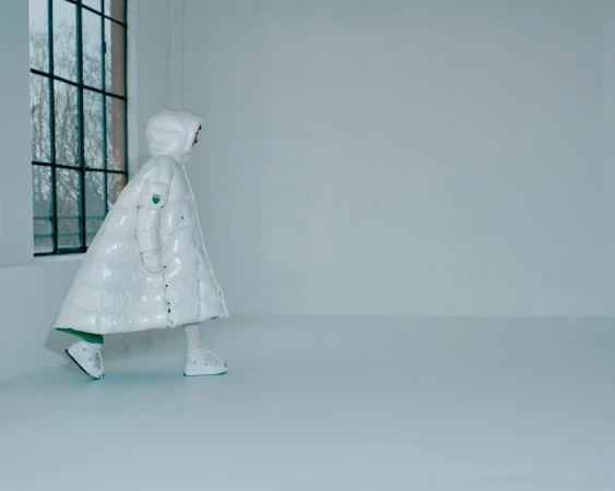 Person in a white puffer coat with hood walking in an empty white room with a window to the left, fashion shot, minimalistic.