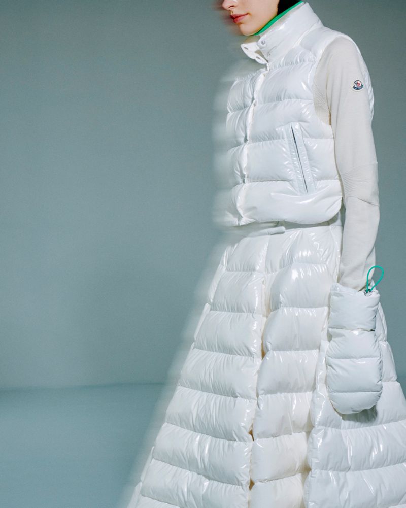 Woman in puffy white Moncler vest and skirt with matching hand warmer in a studio setting on a light gray background.