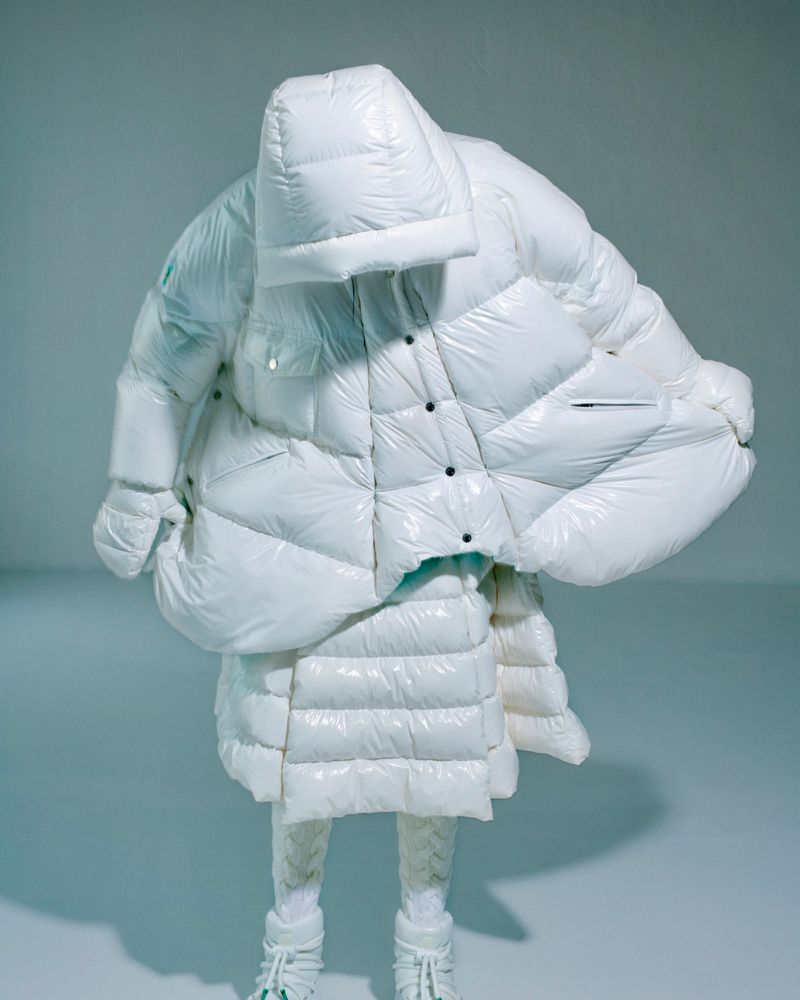 Full-body studio shot of an individual completely enveloped in a white puffy down coat, skirt, gloves, and boots ensemble.
