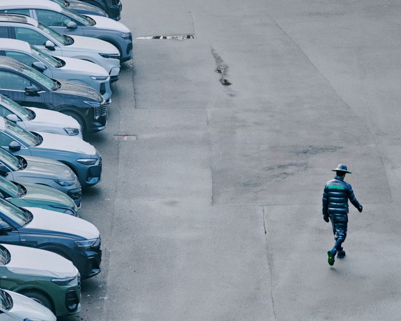 Man walking in a parking lot near parked cars. The man is wearing a hat and dark clothing with bright green accents.