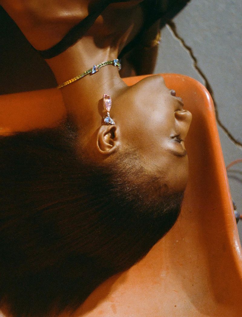 Woman with dark hair reclines in an orange chair wearing jeweled gold choker and earrings. Bright, soft lighting.
