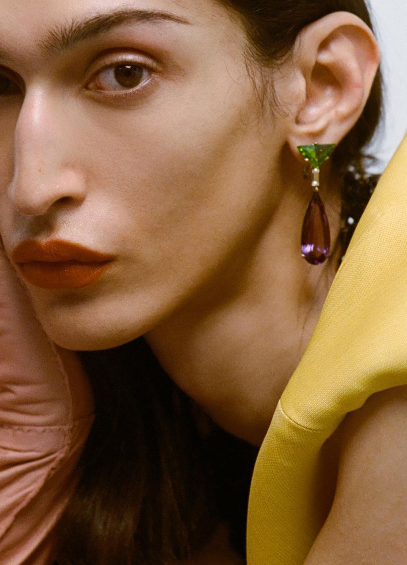 Close-up of a person with dark hair wearing gemstone earrings with yellow fabric draped around their shoulders in a studio setting.