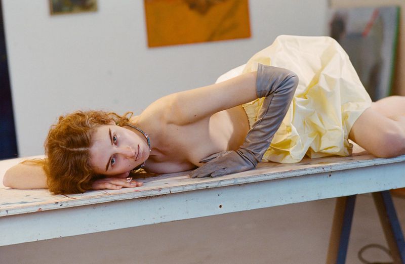 Red-haired woman wearing grey opera gloves and yellow dress lying on a white table in an artist's studio filled with abstract paintings.