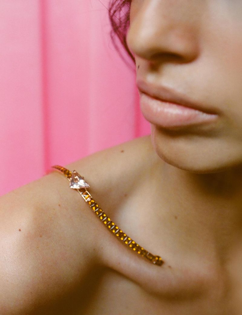Close-up of a woman's shoulder and face with a gold chain necklace draped over her shoulder against a pink curtain backdrop.