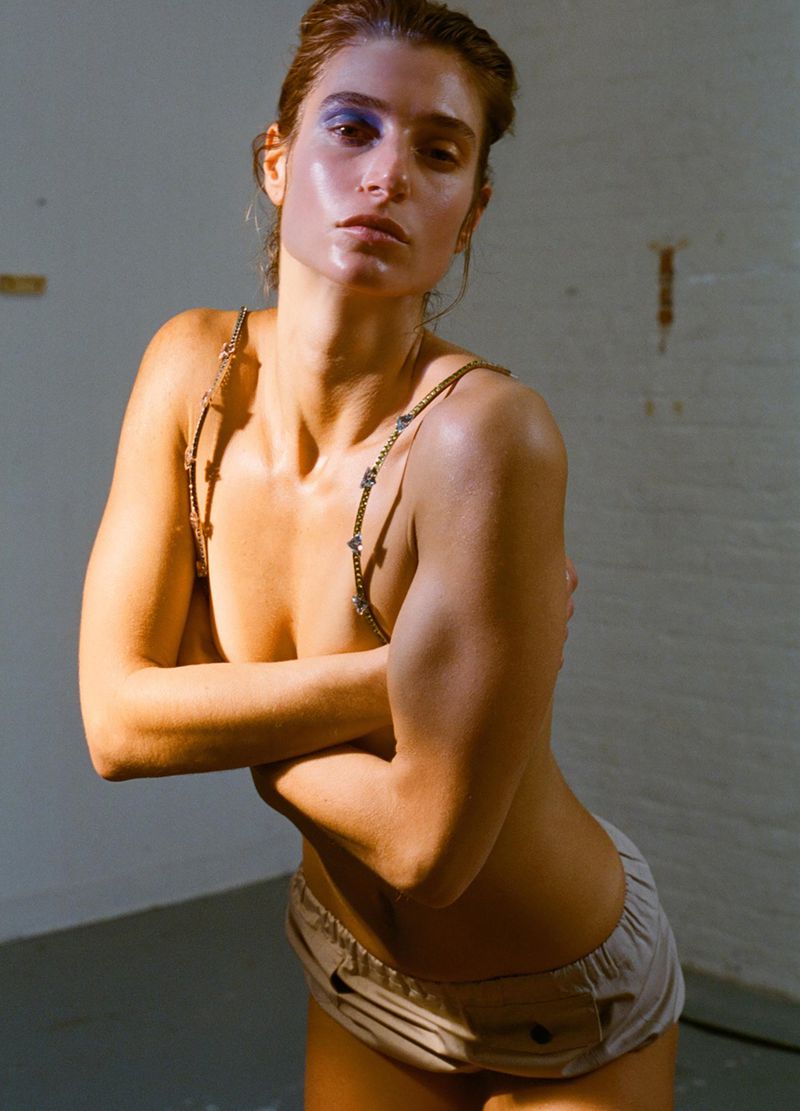 Model with blue eyeshadow and chained straps posing against a neutral wall for an AnOther Magazine editorial by Brianna Capozzi