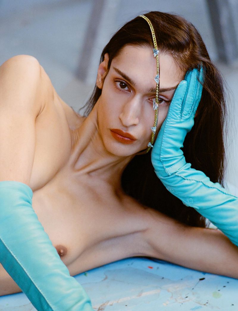 Transgender model poses with long hair, light-blue gloves, and jeweled headpiece on a light-blue surface in a studio fashion editorial.