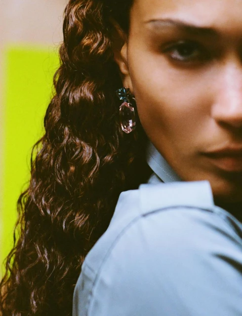 Close-up portrait of a young woman with brown wavy hair, wearing a light blue shirt and jewel earrings against a green and yellow backdrop.