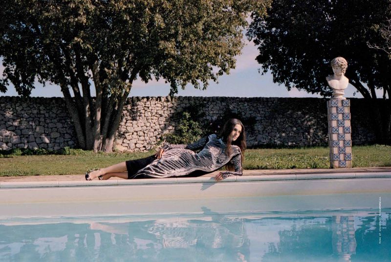 A woman in a patterned dress reclines by a pool, surrounded by lush greenery and a classic bust on a pedestal in a tranquil setting.