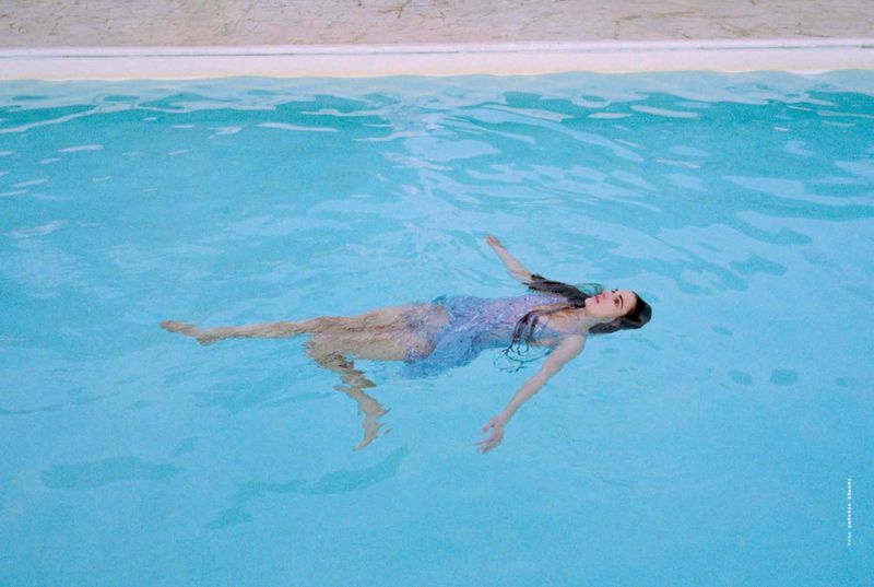 Young woman in a sheer blue dress floats peacefully in a bright blue swimming pool with a white tiled edge, capturing a serene scene.