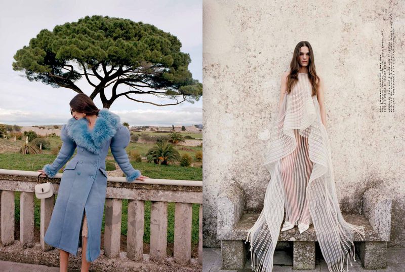 Two fashion models pose outdoors in different outfits. One wears a blue coat with fur trim, the other a white sheer dress.