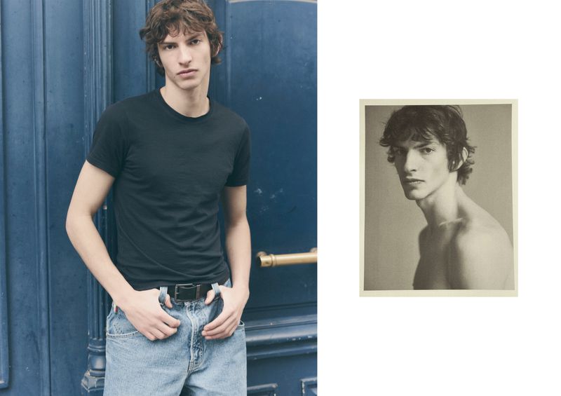Young man wearing a black t-shirt and light blue jeans standing against a dark blue paneled door with an inset black and white portrait.