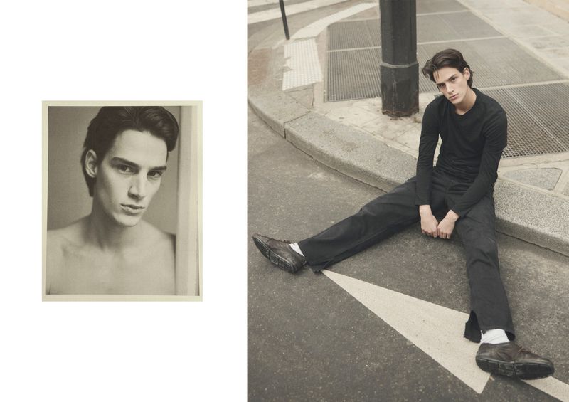Adult male fashion model poses on urban street near curb with arrow, wearing black top and pants, and shoes, photographed with desaturated color.