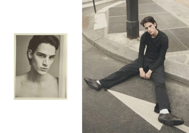 Adult male fashion model poses on urban street near curb with arrow, wearing black top and pants, and shoes, photographed with desaturated color.