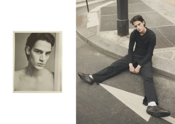 A young male model seated on a paved street next to an inset portrait photograph, wearing a black long-sleeve top and dark trousers.