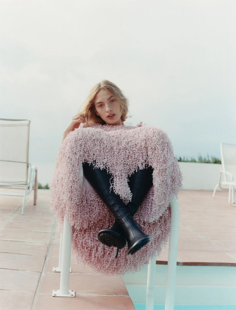 A blonde woman sits by a swimming pool in an oversized pink fuzzy sweater and tall black boots, posing for a fashion editorial.